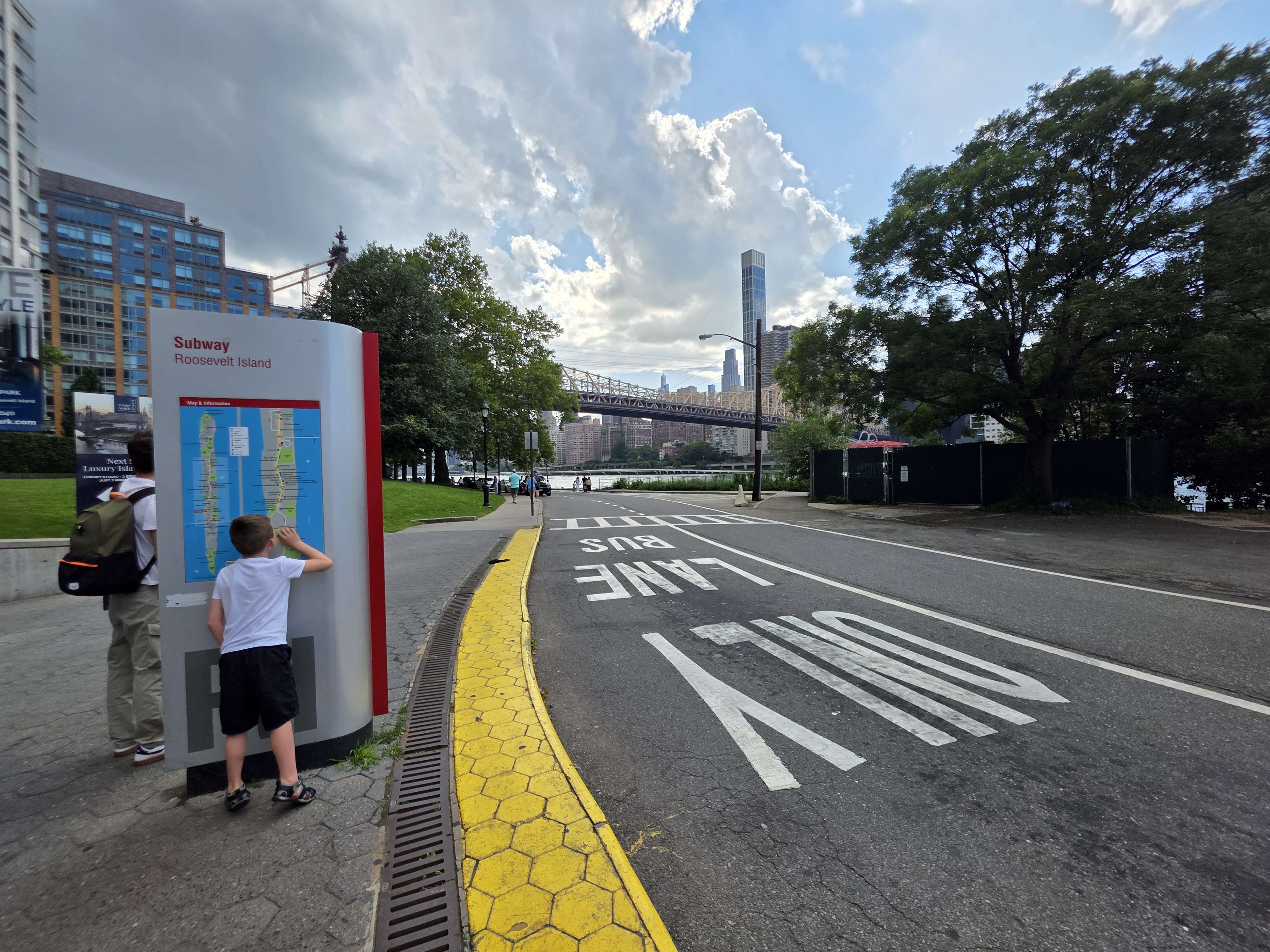 A child at the Roosevelt Island subway map, with the Queensboro Bridge and Manhattan skyline beyond.